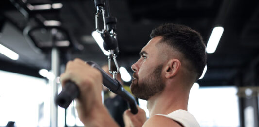 Quer costas mais largas? Veja por que o pulley pronado não pode faltar no seu treino Atleta executando pulley pronado em equipamento profissional de academia - treino de costas
