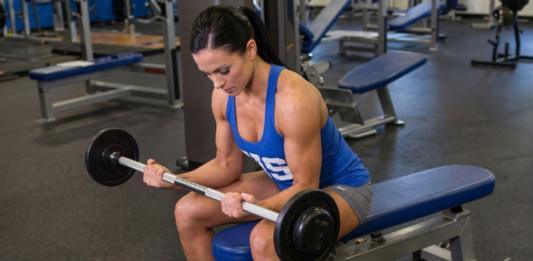 Mulher realiza exercício de rosca de punho com barra sentada em banco na academia, focando no fortalecimento do antebraço.