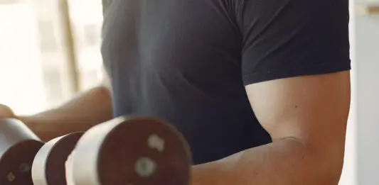 Homem treinando braços com halteres em academia.