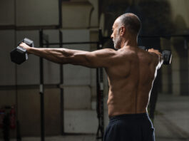 Exercícios para ombros são o divisor entre um treino forte e um ombro lesionado Homem visto de costas realiza elevação lateral com halteres em uma academia, destacando a ativação dos músculos dos ombros e das costas.