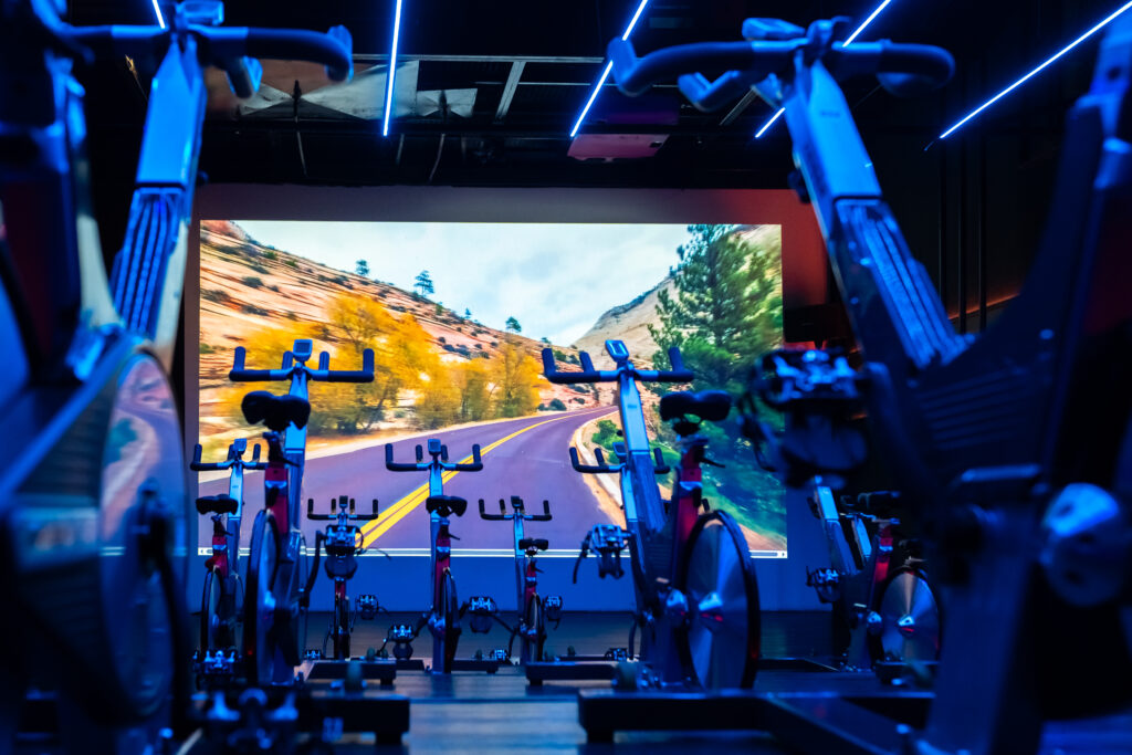 Sala de spinning da Cia Athletica com bicicletas ergométricas alinhadas, iluminação LED azul e painel de projeção imersivo ao fundo exibindo paisagem de estrada em canyon. 