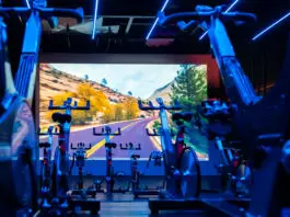Sala de spinning da Cia Athletica com bicicletas ergométricas alinhadas, iluminação LED azul e painel de projeção imersivo ao fundo exibindo paisagem de estrada em canyon.