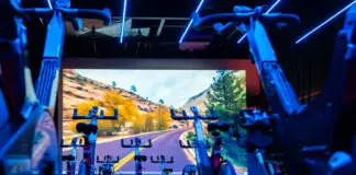 Sala de spinning da Cia Athletica com bicicletas ergométricas alinhadas, iluminação LED azul e painel de projeção imersivo ao fundo exibindo paisagem de estrada em canyon.