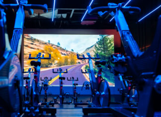 Academia premium: o que realmente muda na experiência, no cuidado e na evolução Sala de spinning da Cia Athletica com bicicletas ergométricas alinhadas, iluminação LED azul e painel de projeção imersivo ao fundo exibindo paisagem de estrada em canyon.