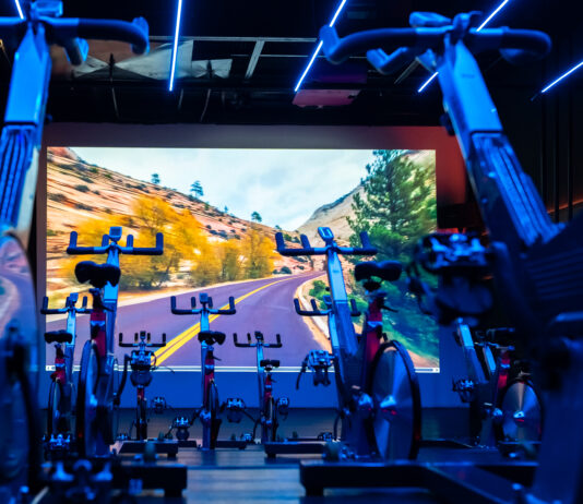 Academia premium: o que realmente muda na experiência, no cuidado e na evolução Sala de spinning da Cia Athletica com bicicletas ergométricas alinhadas, iluminação LED azul e painel de projeção imersivo ao fundo exibindo paisagem de estrada em canyon.