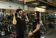 Instrutora acompanha aluno durante exercício com halteres, corrigindo execução em sala de musculação.
