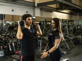 Instrutora acompanha aluno durante exercício com halteres, corrigindo execução em sala de musculação.