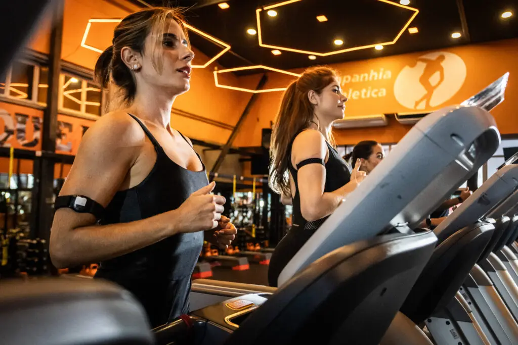 Mulheres correndo em esteiras em academia moderna com iluminação quente, durante treino cardiovascular focado em bem-estar e redução do estresse.