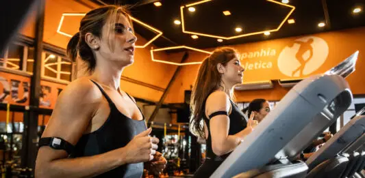 Mulheres correndo em esteiras em academia moderna com iluminação quente, durante treino cardiovascular focado em bem-estar e redução do estresse.