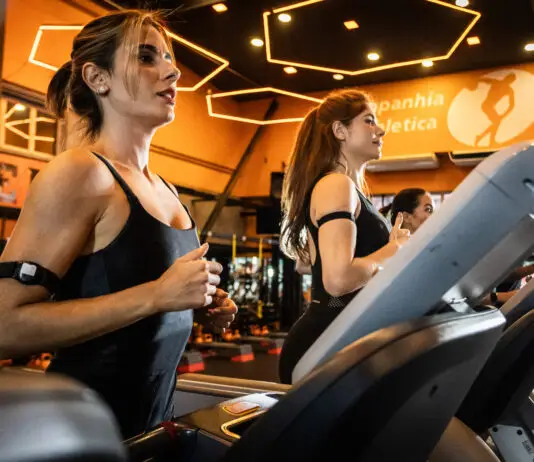 Mulheres correndo em esteiras em academia moderna com iluminação quente, durante treino cardiovascular focado em bem-estar e redução do estresse.