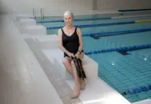 Idosa sorridente sentada na borda de uma piscina indoor, usando maiô preto e touca de natação, segurando nadadeiras, em ambiente de hidroginástica e atividades aquáticas para idosos.