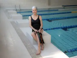 Idosa sorridente sentada na borda de uma piscina indoor, usando maiô preto e touca de natação, segurando nadadeiras, em ambiente de hidroginástica e atividades aquáticas para idosos.