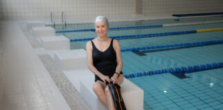 Idosa sorridente sentada na borda de uma piscina indoor, usando maiô preto e touca de natação, segurando nadadeiras, em ambiente de hidroginástica e atividades aquáticas para idosos.