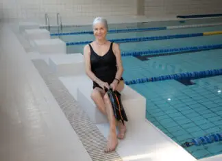 Idosa sorridente sentada na borda de uma piscina indoor, usando maiô preto e touca de natação, segurando nadadeiras, em ambiente de hidroginástica e atividades aquáticas para idosos.