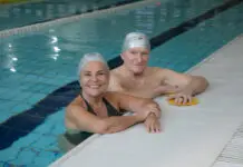 Casal de idosos sorrindo dentro de uma piscina coberta da Cia Athletica, usando toucas de natação durante atividade do Programa Platinum