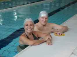 Casal de idosos sorrindo dentro de uma piscina coberta da Cia Athletica, usando toucas de natação durante atividade do Programa Platinum