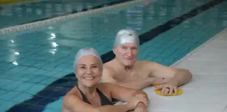 Casal de idosos sorrindo dentro de uma piscina coberta da Cia Athletica, usando toucas de natação durante atividade do Programa Platinum