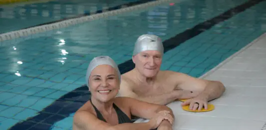 Casal de idosos sorrindo dentro de uma piscina coberta da Cia Athletica, usando toucas de natação durante atividade do Programa Platinum