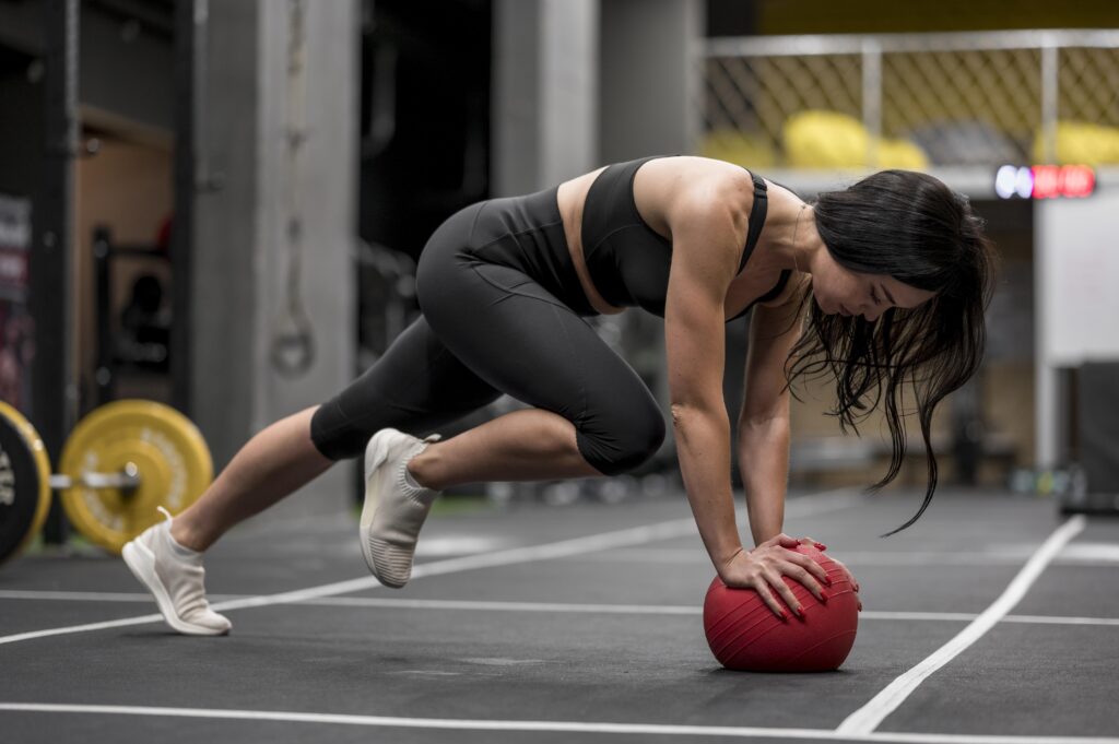 Mulher realiza exercício em posição de prancha com apoio sobre uma bola medicinal durante treino funcional de alta intensidade, representando o HIIT e o condicionamento cardiovascular.