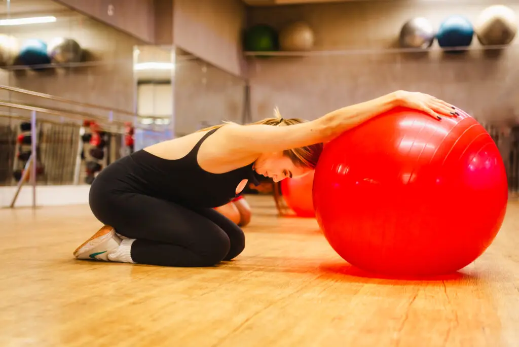 Mulher ajoelhada em uma sala de academia realiza um alongamento com os braços apoiados sobre uma grande bola vermelha de exercícios.
