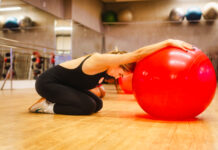 Mulher ajoelhada em uma sala de academia realiza um alongamento com os braços apoiados sobre uma grande bola vermelha de exercícios.
