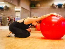 Mulher ajoelhada em uma sala de academia realiza um alongamento com os braços apoiados sobre uma grande bola vermelha de exercícios.
