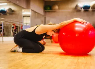 Mulher ajoelhada em uma sala de academia realiza um alongamento com os braços apoiados sobre uma grande bola vermelha de exercícios.