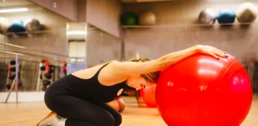Composição corporal: o que é e por que a balança não conta toda a história Mulher ajoelhada em uma sala de academia realiza um alongamento com os braços apoiados sobre uma grande bola vermelha de exercícios.