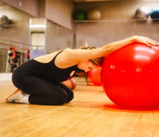 Mulher ajoelhada em uma sala de academia realiza um alongamento com os braços apoiados sobre uma grande bola vermelha de exercícios.