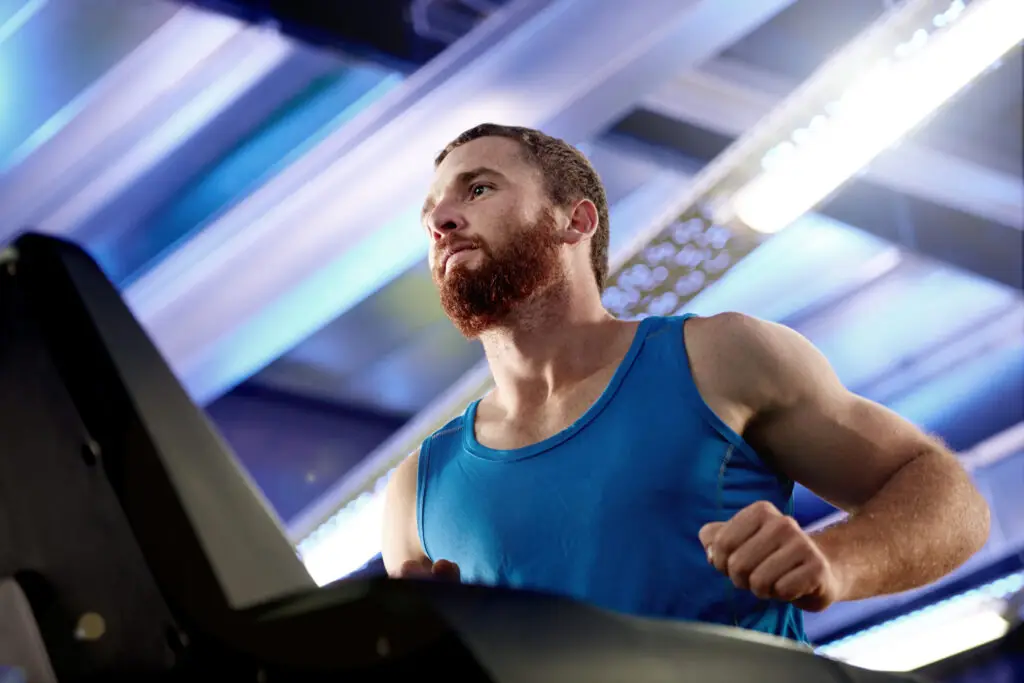 Homem com camiseta azul corre em uma esteira dentro de uma academia, em ambiente iluminado por luzes brancas e azuladas.
