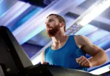 Homem com camiseta azul corre em uma esteira dentro de uma academia, em ambiente iluminado por luzes brancas e azuladas.