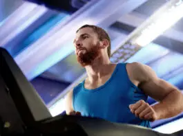 Homem com camiseta azul corre em uma esteira dentro de uma academia, em ambiente iluminado por luzes brancas e azuladas.