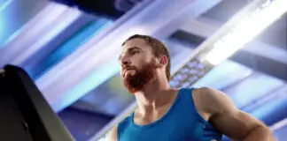 Homem com camiseta azul corre em uma esteira dentro de uma academia, em ambiente iluminado por luzes brancas e azuladas.