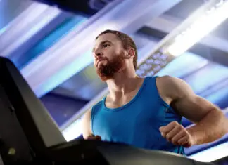 Quanto tempo de cardio para emagrecer? Mais minutos nem sempre significam mais resultado Homem com camiseta azul corre em uma esteira dentro de uma academia, em ambiente iluminado por luzes brancas e azuladas.