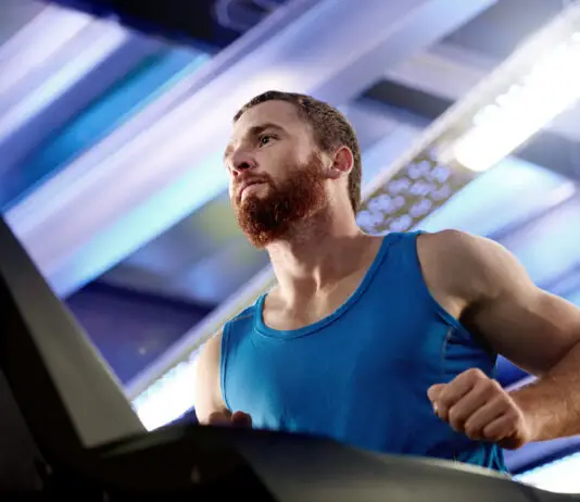 Quanto tempo de cardio para emagrecer? Mais minutos nem sempre significam mais resultado Homem com camiseta azul corre em uma esteira dentro de uma academia, em ambiente iluminado por luzes brancas e azuladas.